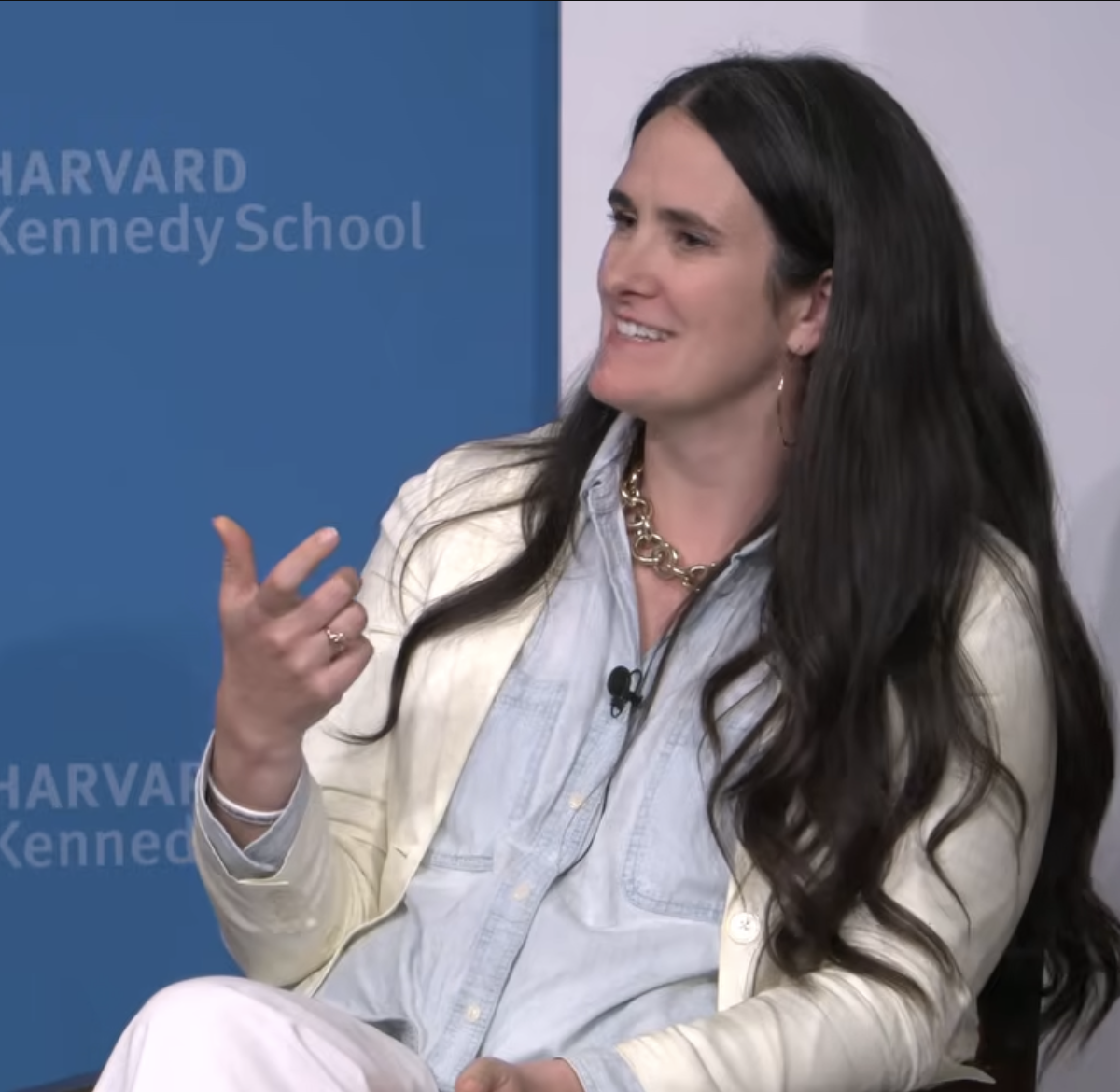 Micaela, a white woman with long, wavy, brown hair, wearing a cream-colored jacket, a blue button-down shirt, and a statement necklace, sits and talks. She is gesturing with her hand and has a microphone clipped to her shirt. Behind her, a blue backdrop with Harvard Kennedy School’s logo patterned on it is visible.